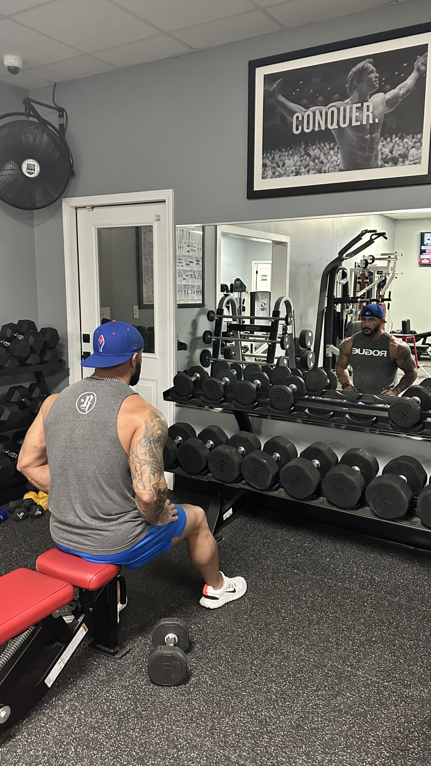 Ben Manville seated at the dumbbell area, reflected in the gym mirror
